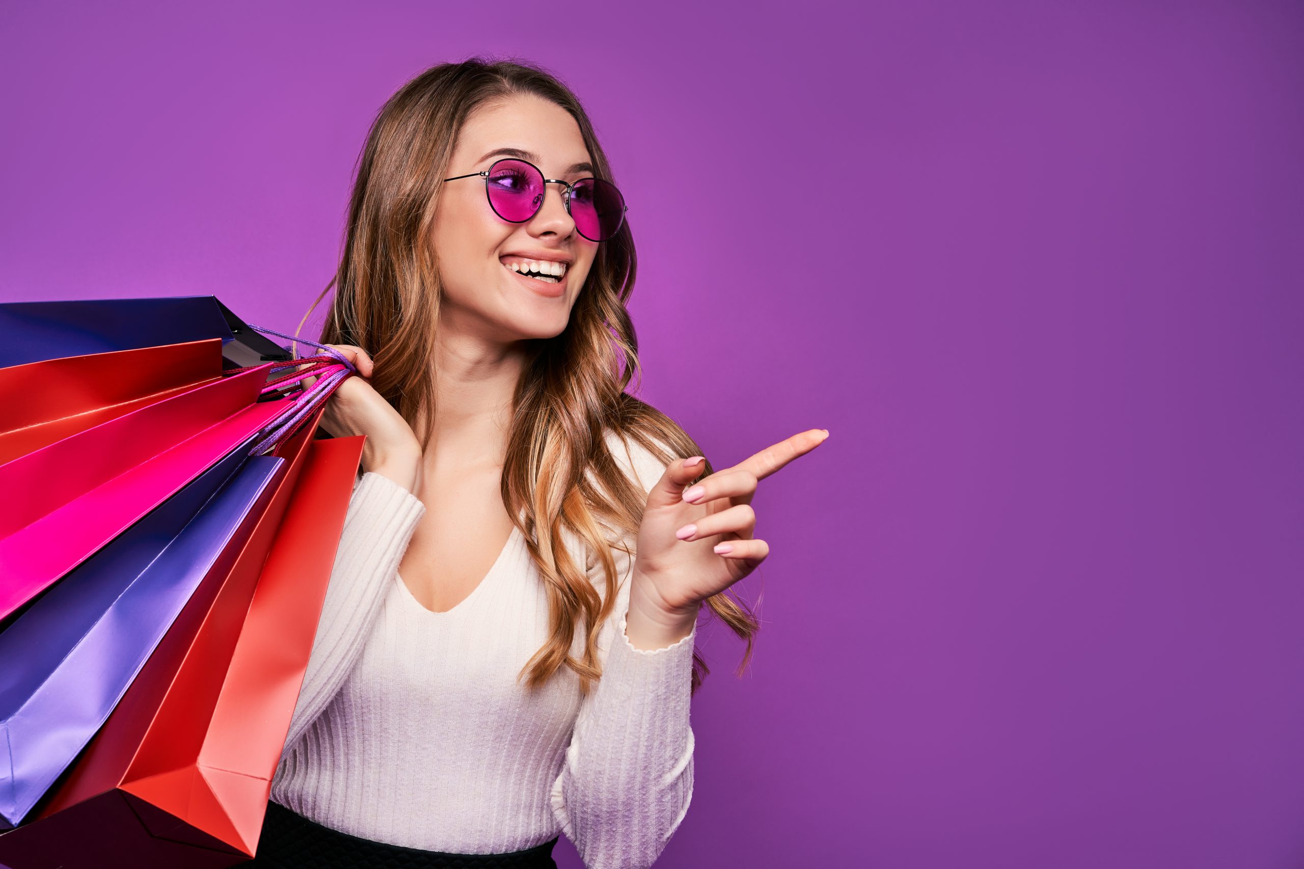 Beautiful smiling young blonde girl pointing in sunglasses holding shopping bags and credit card on a pink background, copy space.