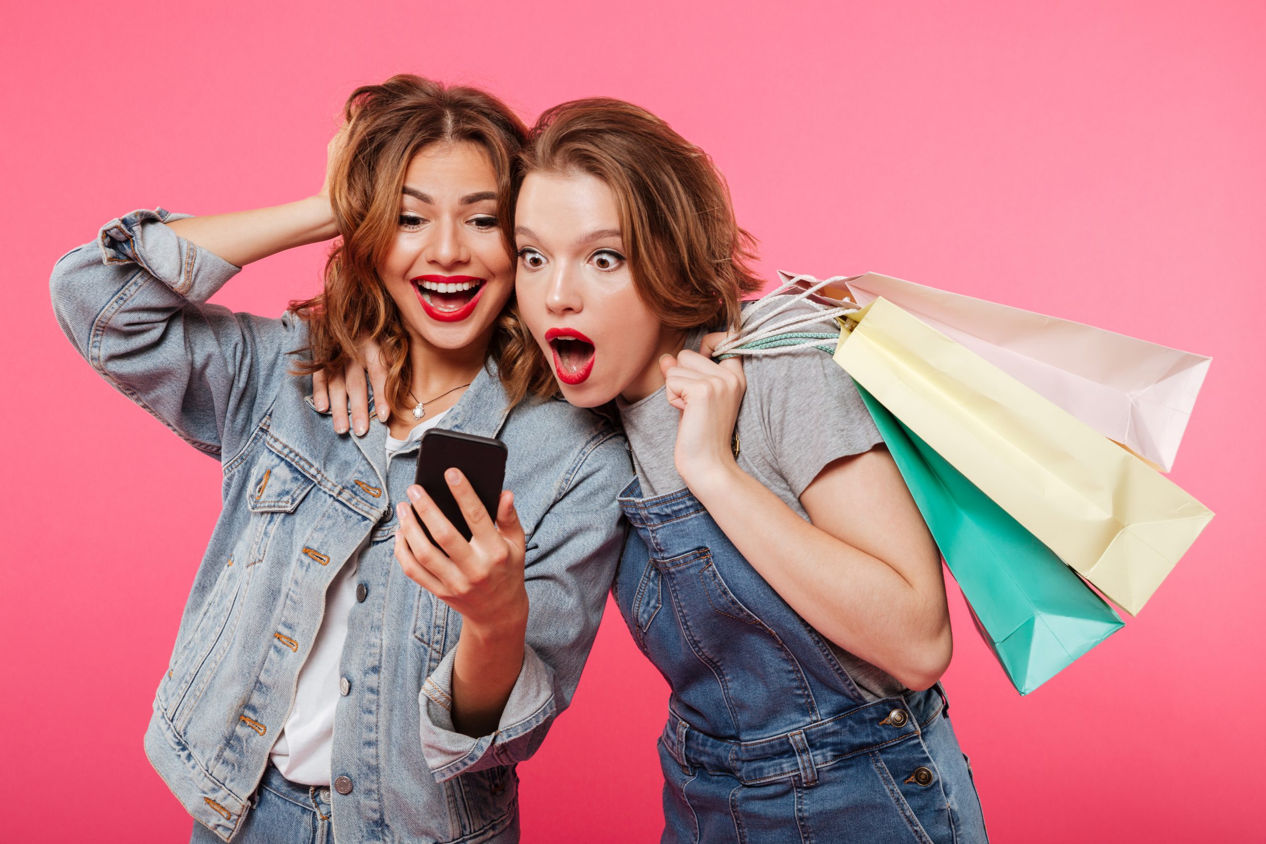Image of shocked two women friends standing isolated over pink background. Looking aside holding shopping bags using mobile phone.
