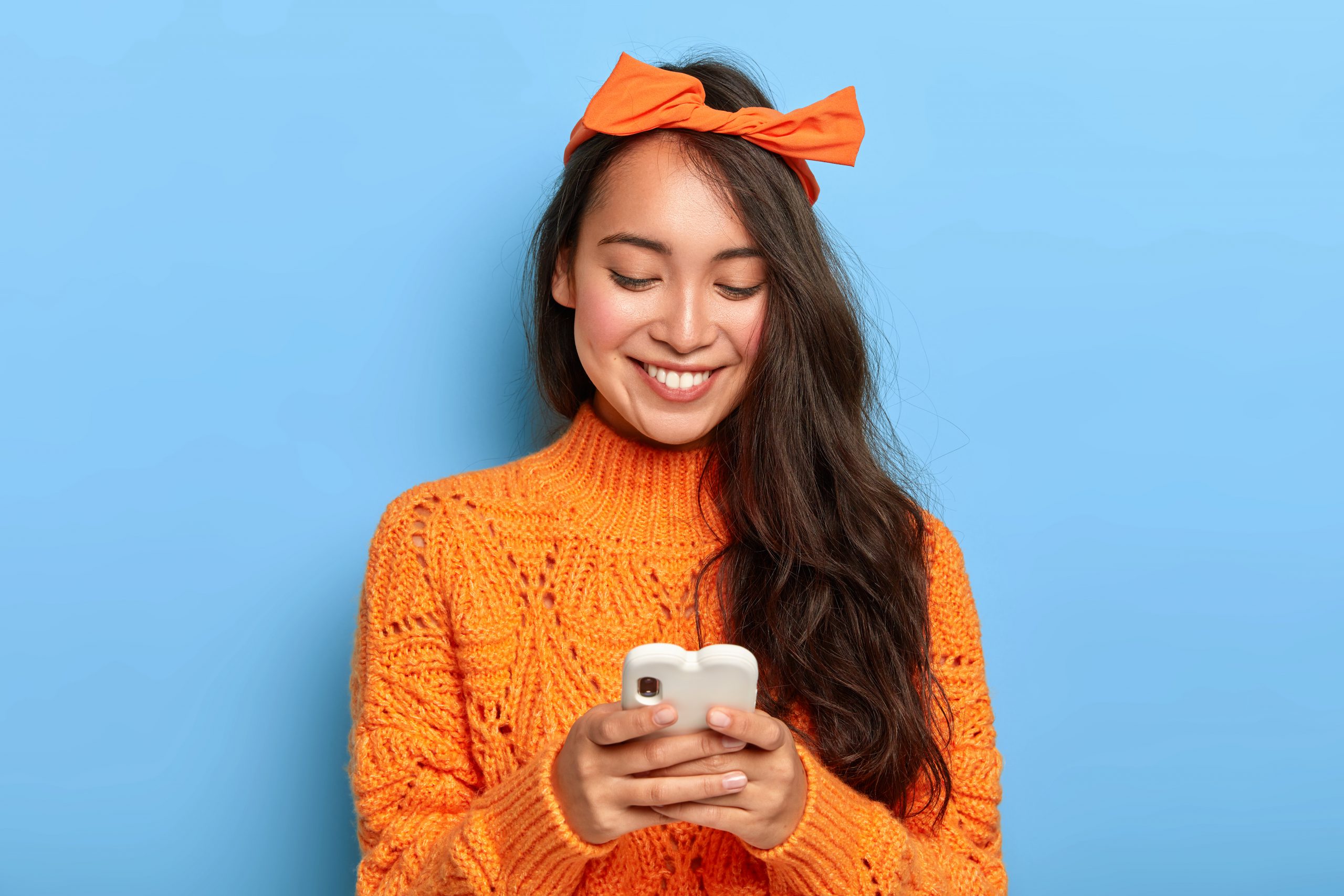 Photo of stylish brunette millennial girl busy checking her email box, holds mobile phone, wears orange headband tied in bow, warm sweater, connected to wireless internet, gets pleasant message