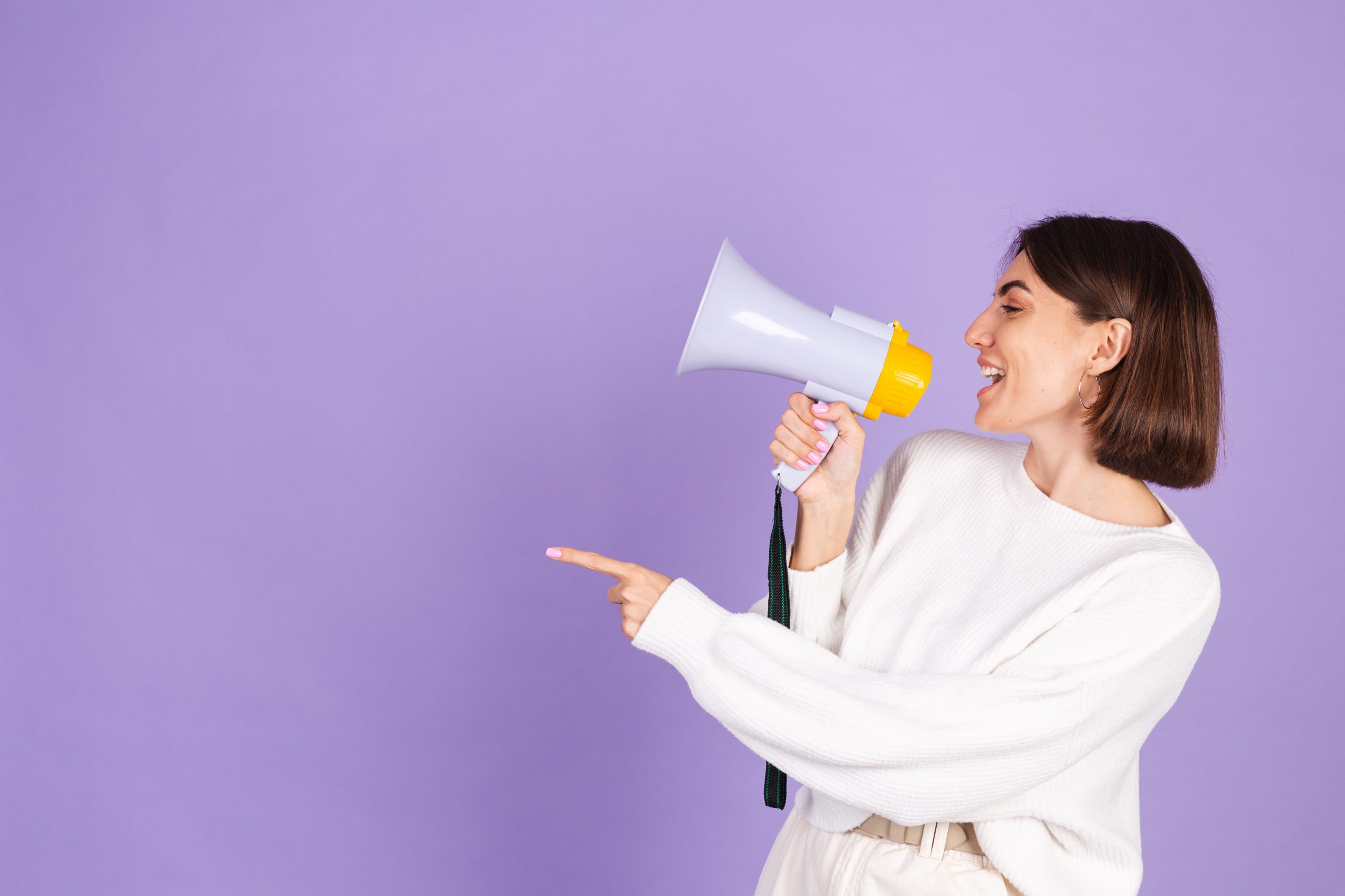 Young brunette in white casual sweater isolated on purple background happy screaming in megaphone copy space point left