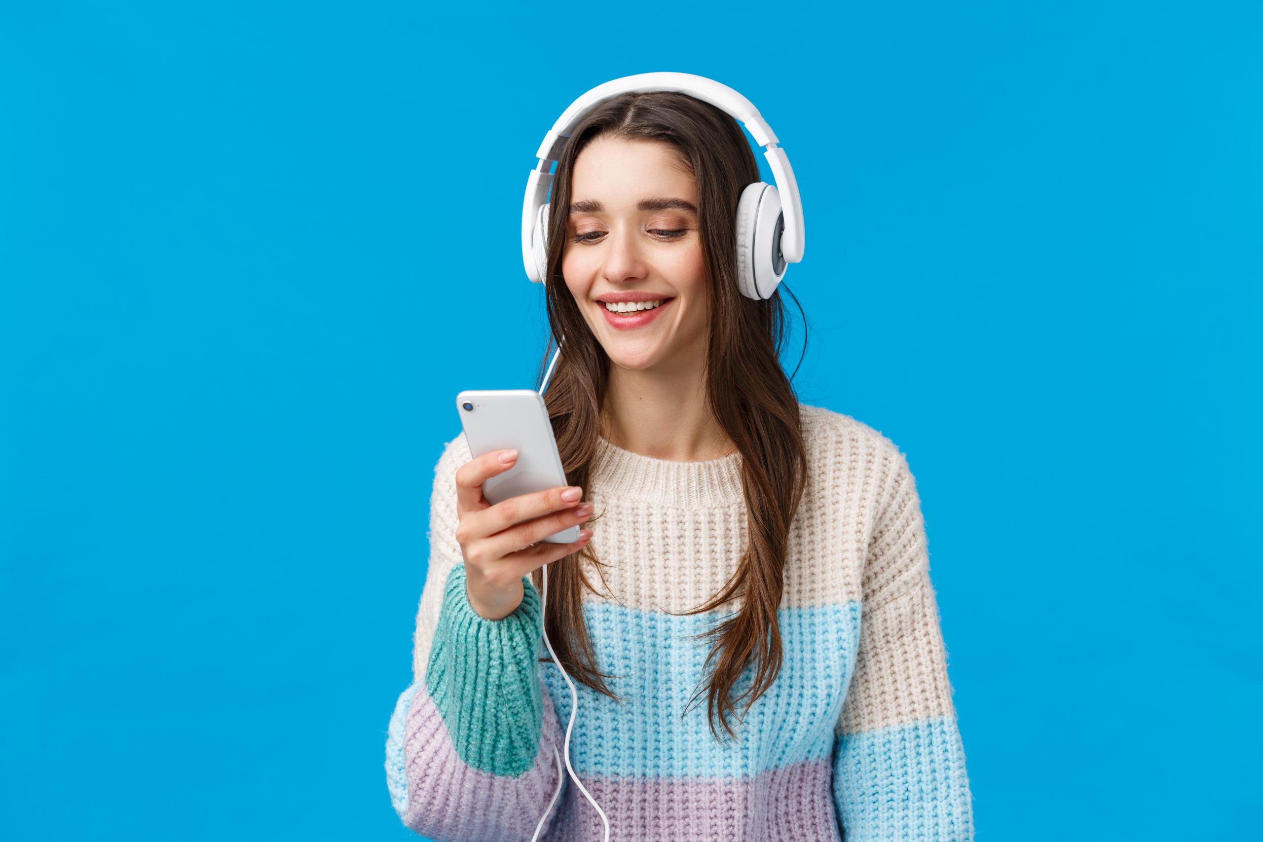 Technology, millennials and lifestyle concept. Carefree cute brunette female student put on headphones, plug-in smartphone picking song and smiling, standing blue background make playlist for study.