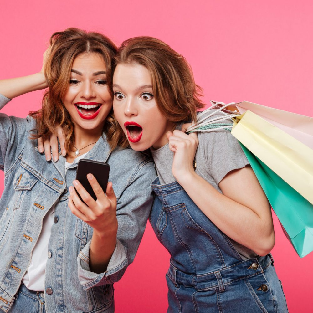 Image of shocked two women friends standing isolated over pink background. Looking aside holding shopping bags using mobile phone.